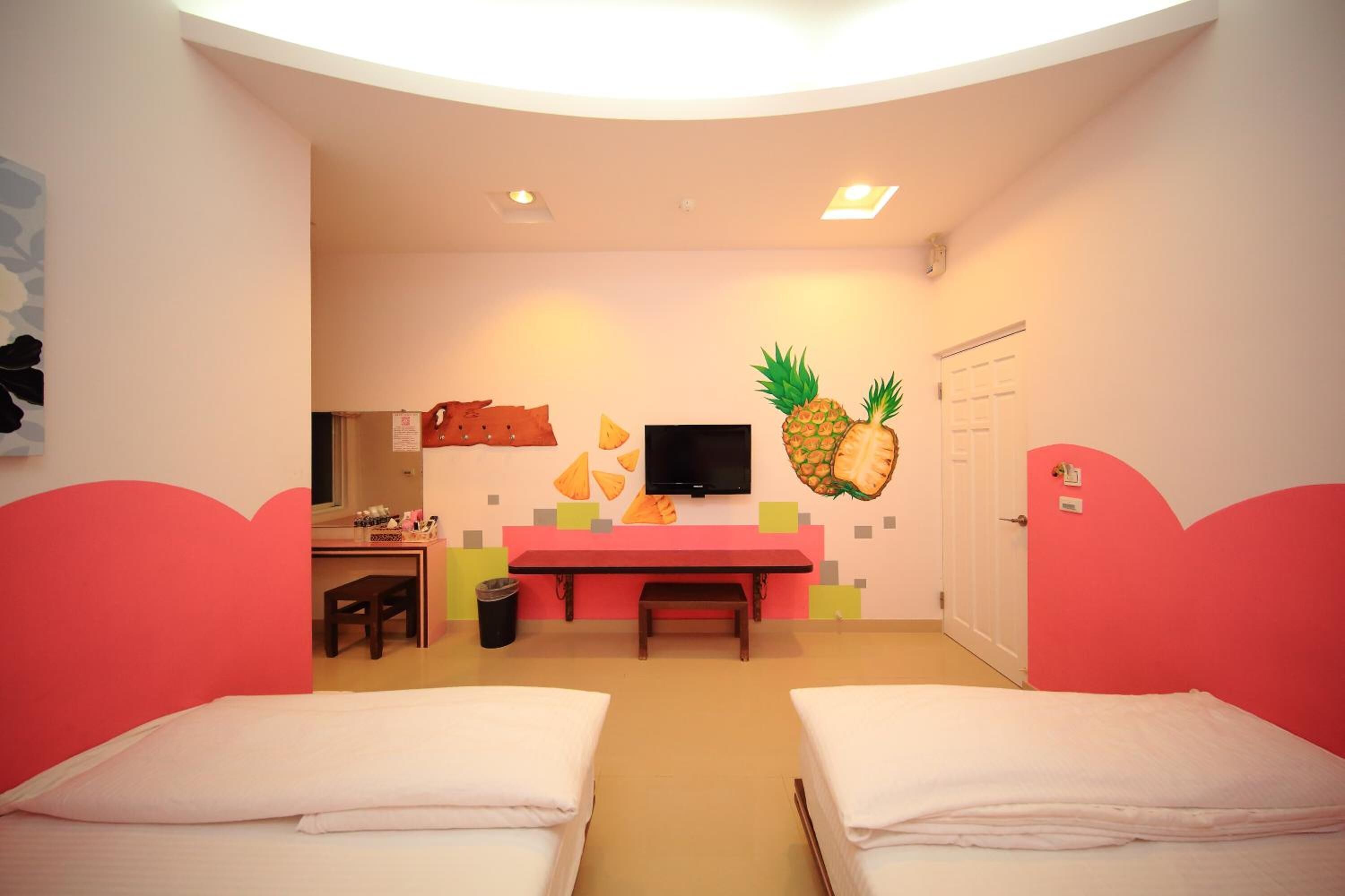Quarto com duas camas, uma escrivaninha, uma cadeira, televisão e um painel decorado com abacaxi.