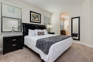 A bedroom with a large bed, two bedside tables, a nightstand, a mirror, and a window with blinds.