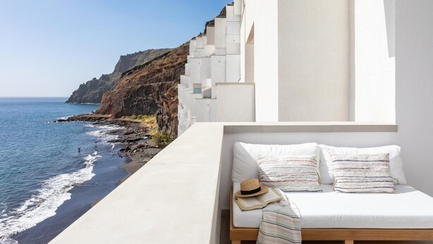Un balcón con un sofá blanco, un sombrero y un libro abierto, con vistas a un litoral rocoso y al mar.