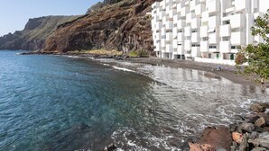Un edificio frente al mar, con un acantilado rocoso de fondo, agua azul cristalina y una playa arenosa.