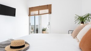 Un dormitorio moderno con una cama grande, un televisor de pantalla plana en la pared, una puerta corrediza de vidrio que da a un balcón y una planta en maceta a la derecha.