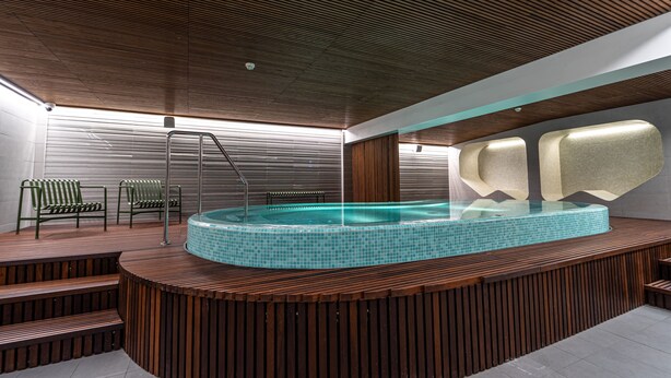 Ein moderner Poolbereich im Innenbereich mit Holzterrasse, einem durch eine Glasscheibe geschützten Whirlpool und einer Metalltreppe.