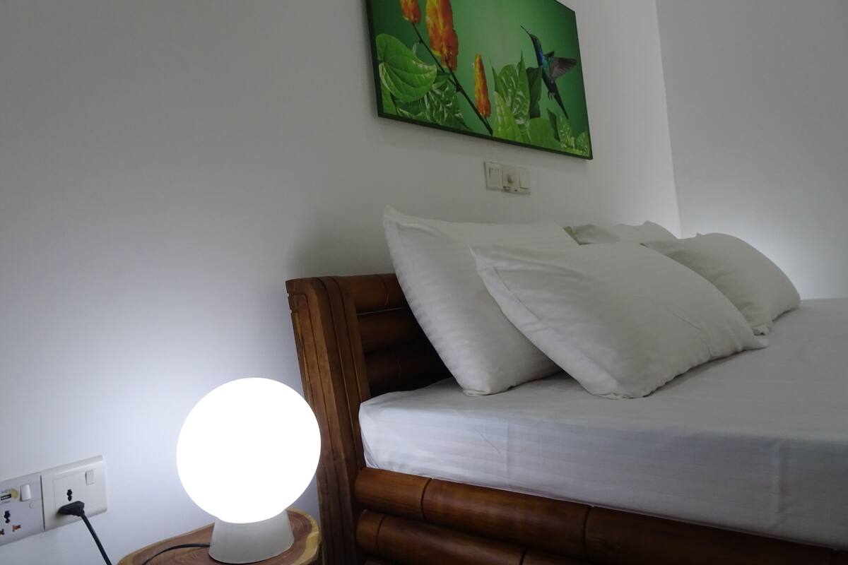 A neatly made bed with white linens and pillows, a bedside lamp on a wooden nightstand, and a painting of birds and flowers on the wall.