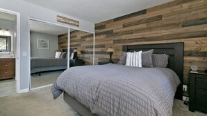 A bedroom with a large bed, a wooden accent wall, a mirror, and a nightstand with a lamp. - Modern Condo with Shared Hot Tub! Short Walk to Village! (Mammoth Lakes)