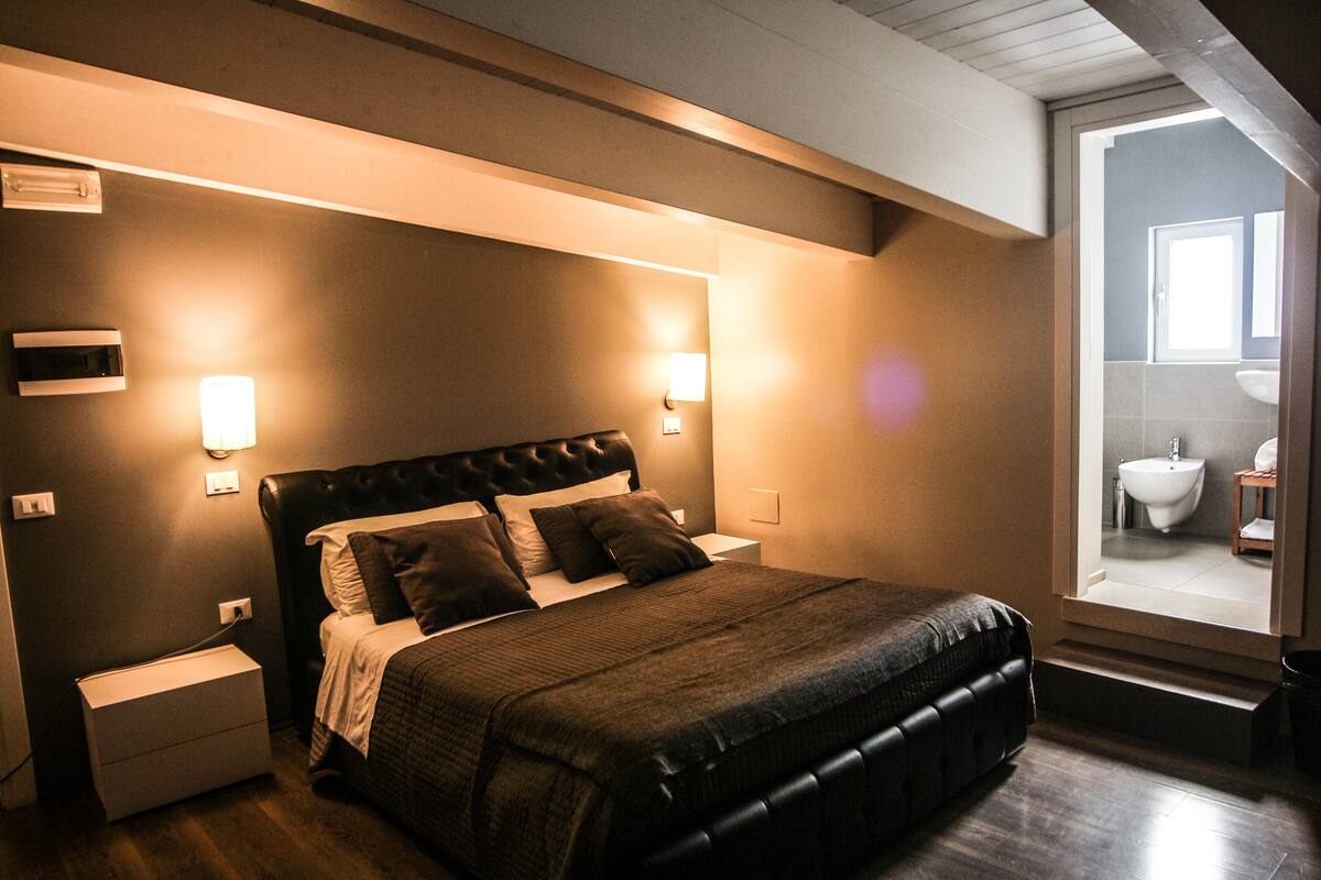 Une chambre à coucher avec un grand lit, des lampes de chevet, une table de nuit et une vue sur la salle de bain par une porte ouverte.