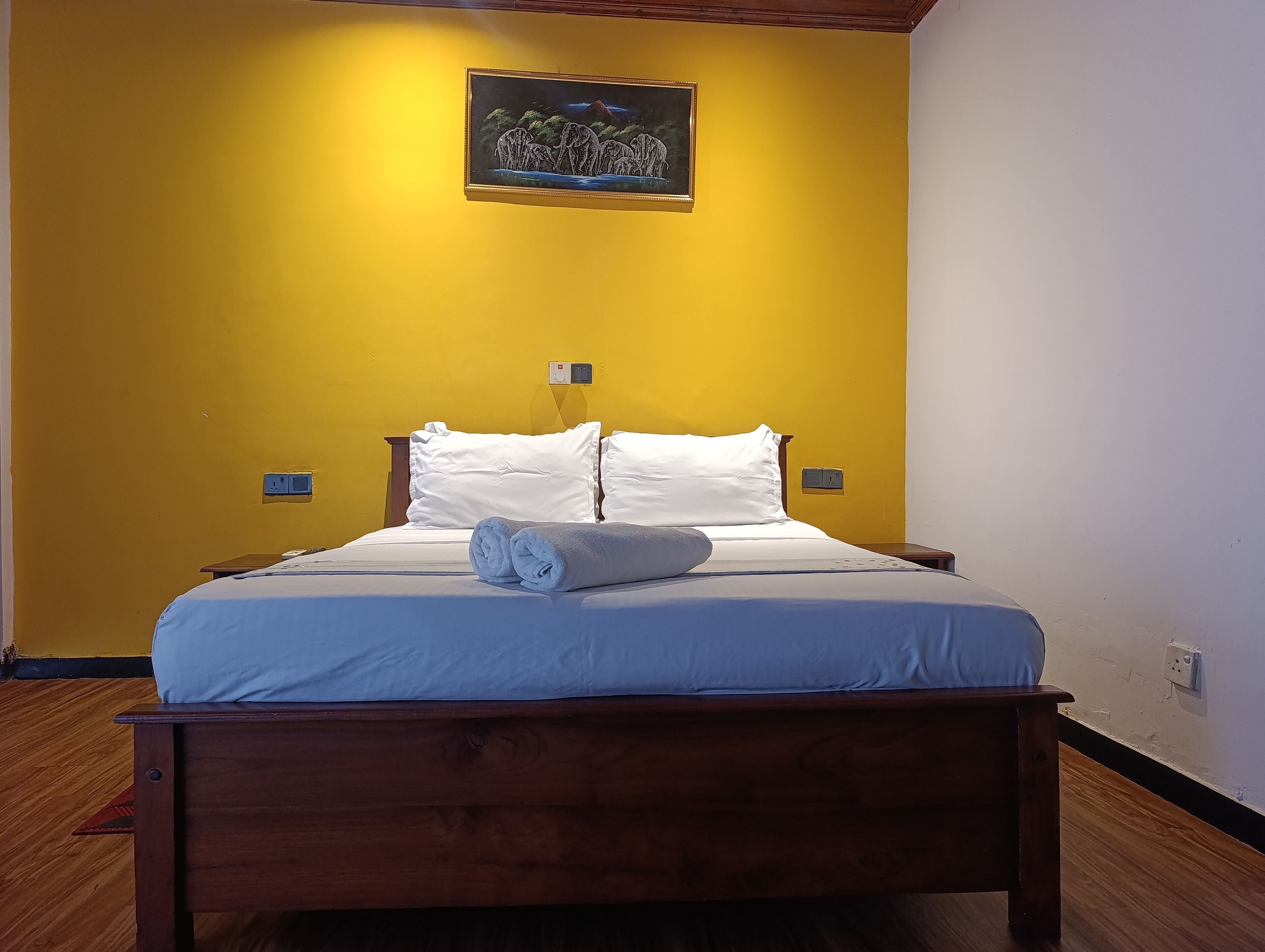 A bed with white linens and a rolled towel, a nightstand, and a wall-mounted light switch.