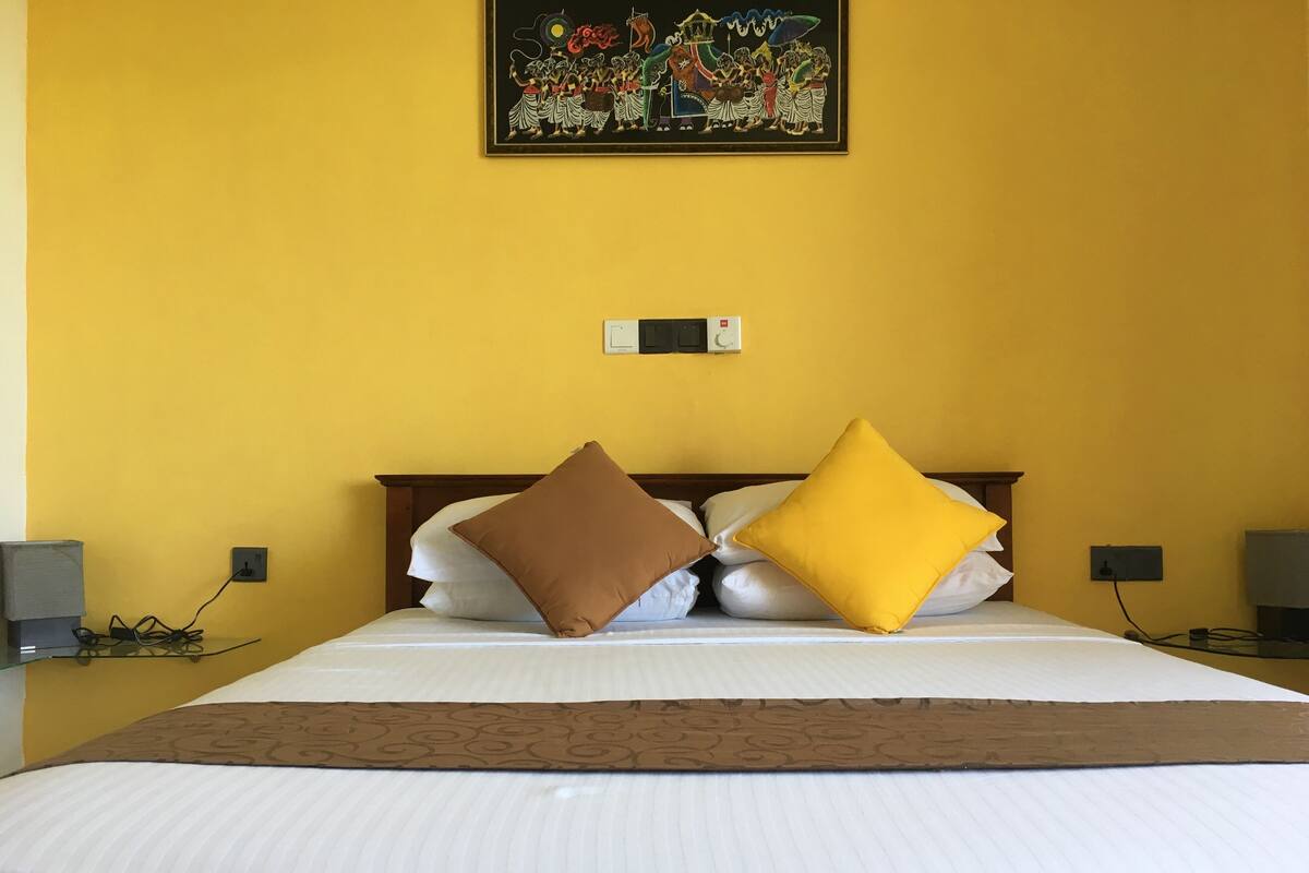A hotel room with a bed, yellow walls, a painting, and a power outlet.