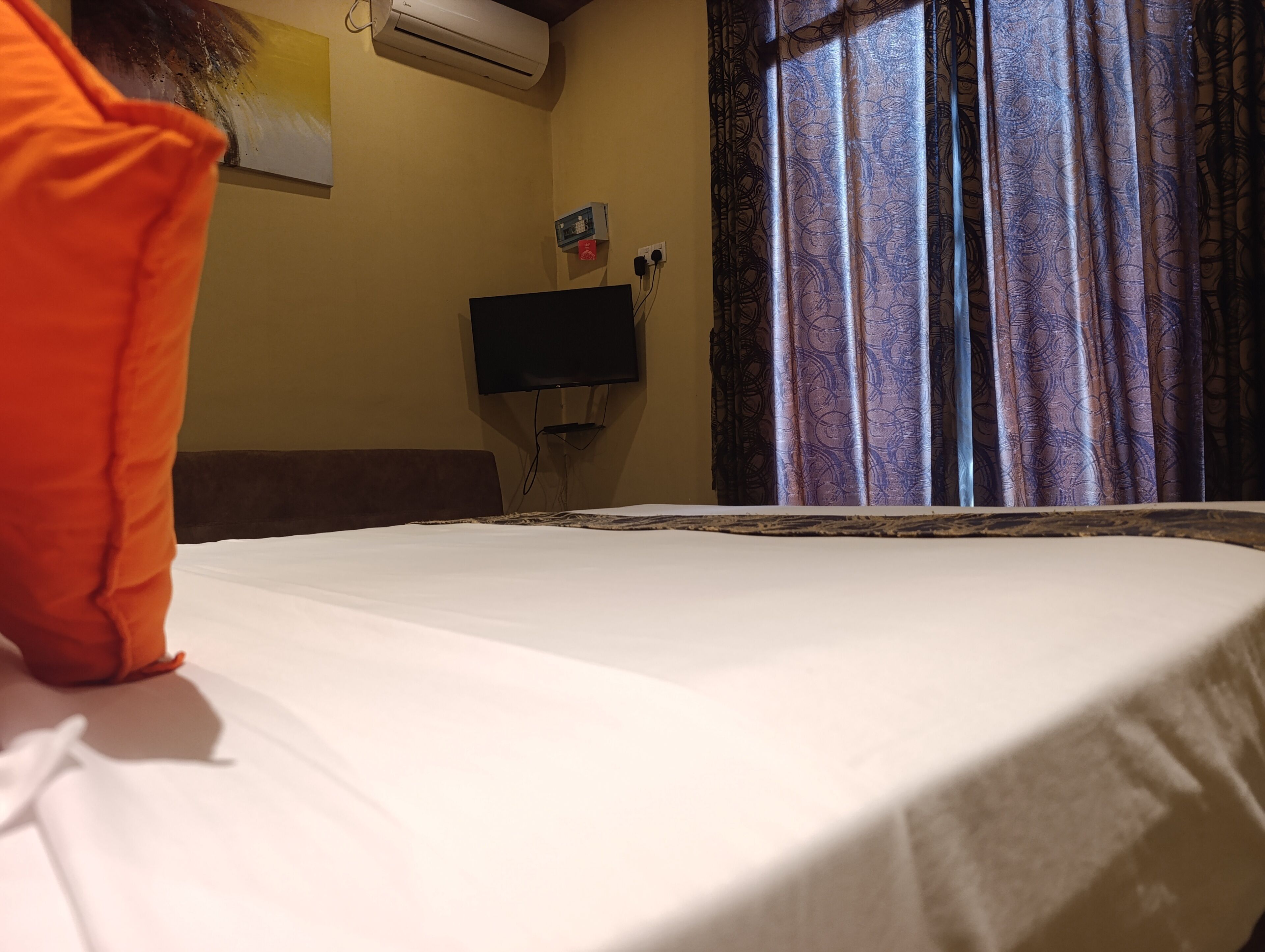 Un lit avec un drap blanc, un téléviseur fixé au mur et une fenêtre ornée de rideaux à motifs.