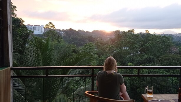 A balcony with a view of a forested landscape and a building in the distance. There is a table with a bottle and a glass. - Villa Forest View (Kandy)