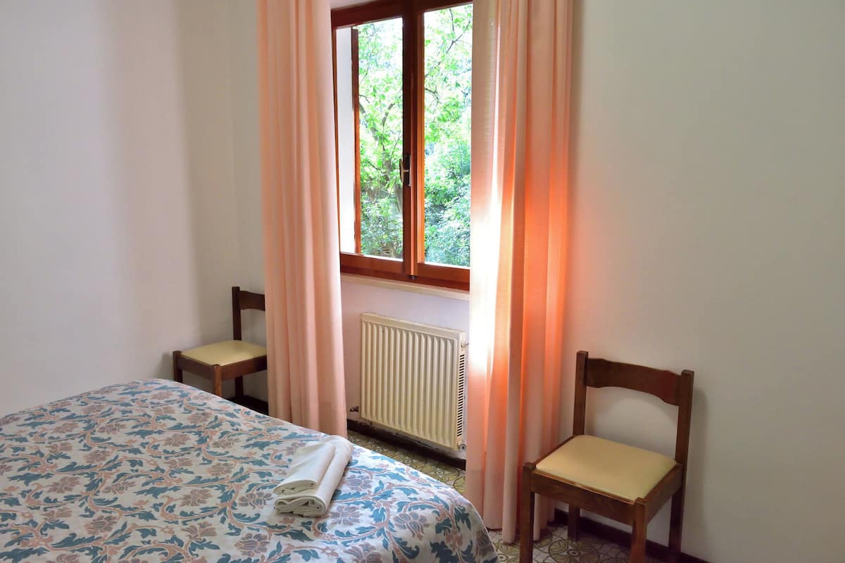 Un dormitorio con una cama, dos sillas, un ventanal con cortinas y un radiador.