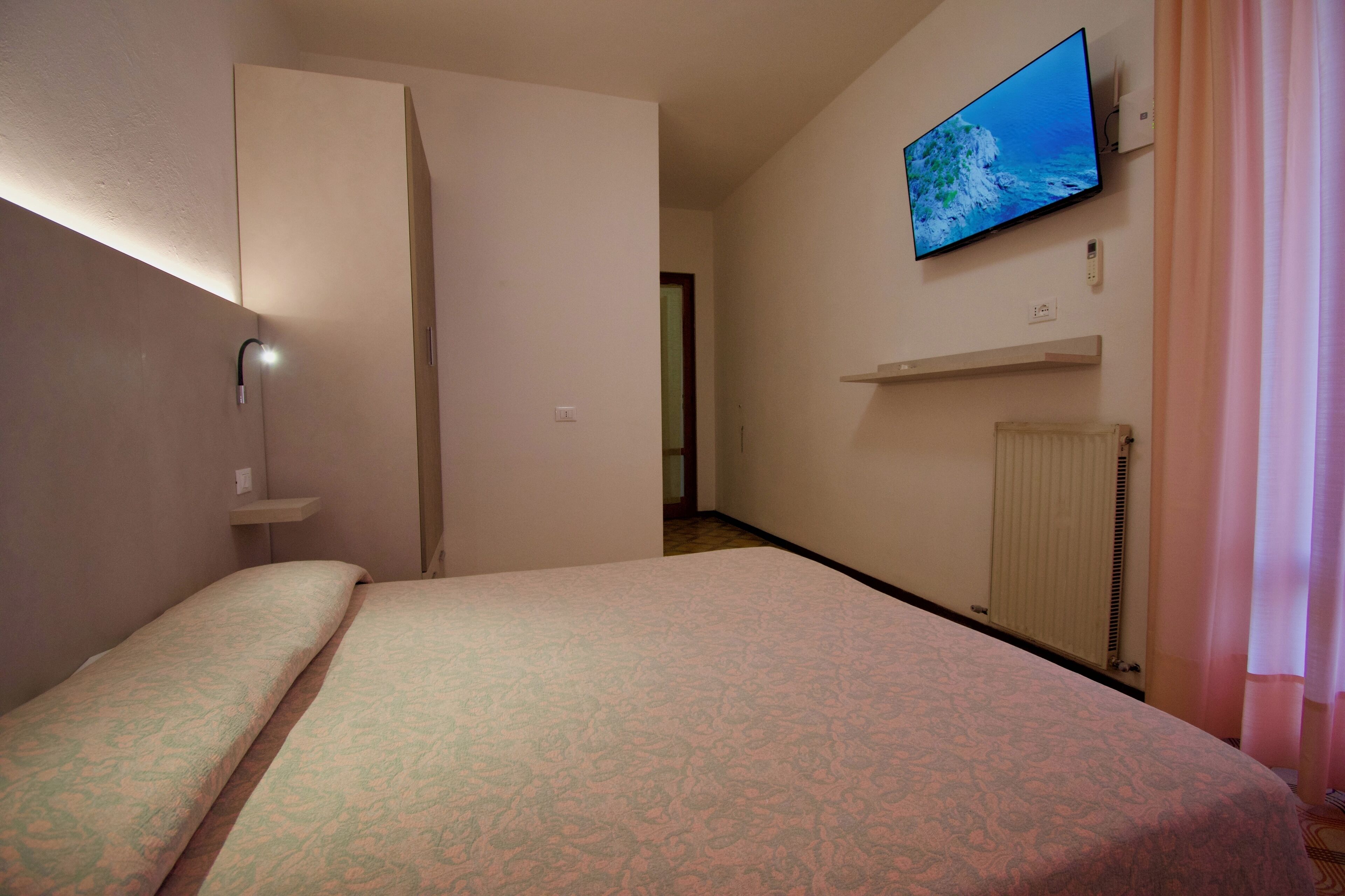 Habitación de hotel con una cama, un televisor montado en la pared, un radiador y una puerta que da a otra habitación.