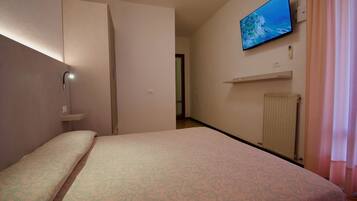 Habitación de hotel con una cama, un televisor montado en la pared, un radiador y una puerta que da a otra habitación.