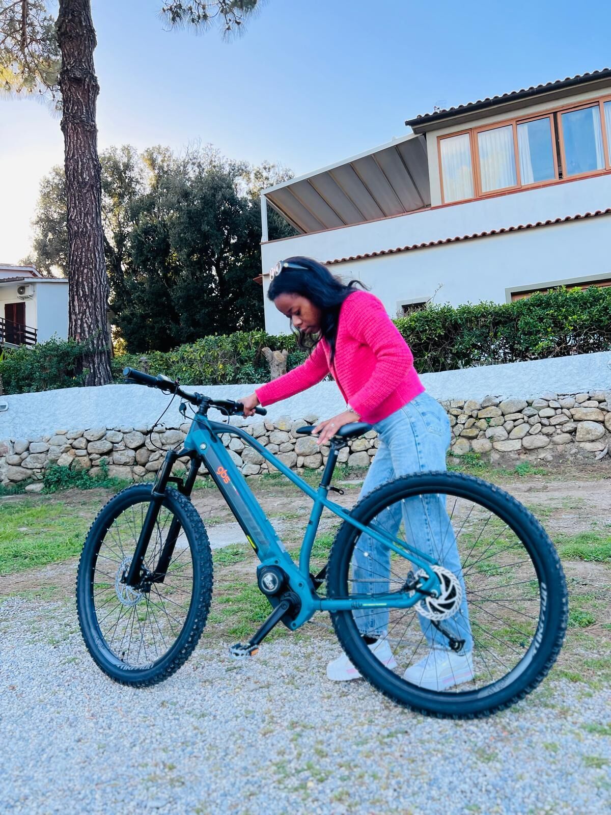 Eine Person justiert ein türkisfarbenes Fahrrad mit Federgabel und Scheibenbremsen, stehend auf einem Schotterweg, im Hintergrund eine Steinmauer und ein modernes Gebäude.