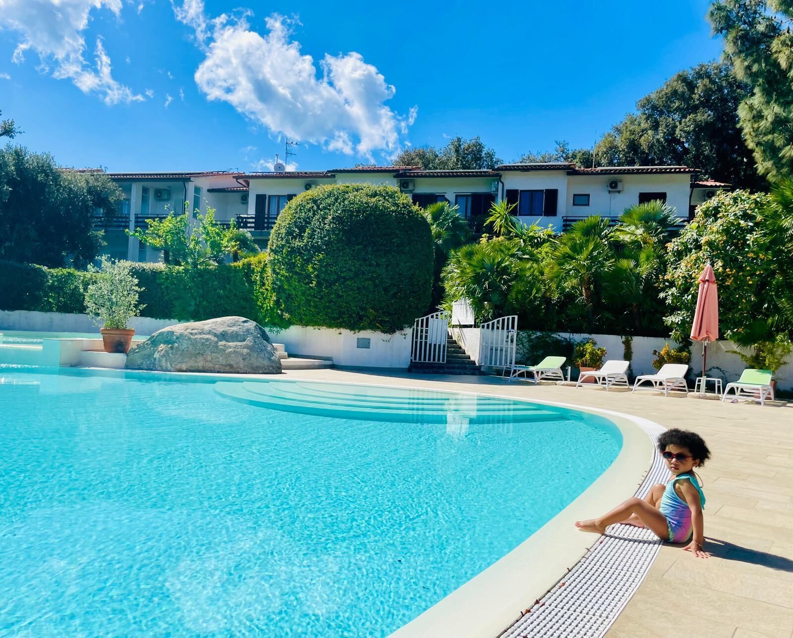 En swimmingpool med et barn, der sidder på kanten, omgivet af liggestole og plantekasser.
