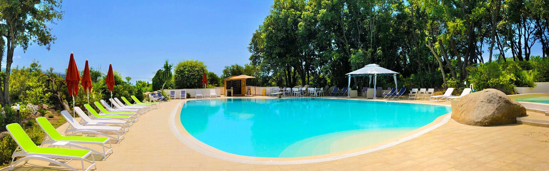 Una piscina con lettini e ombrelloni, circondata da alberi e un padiglione.