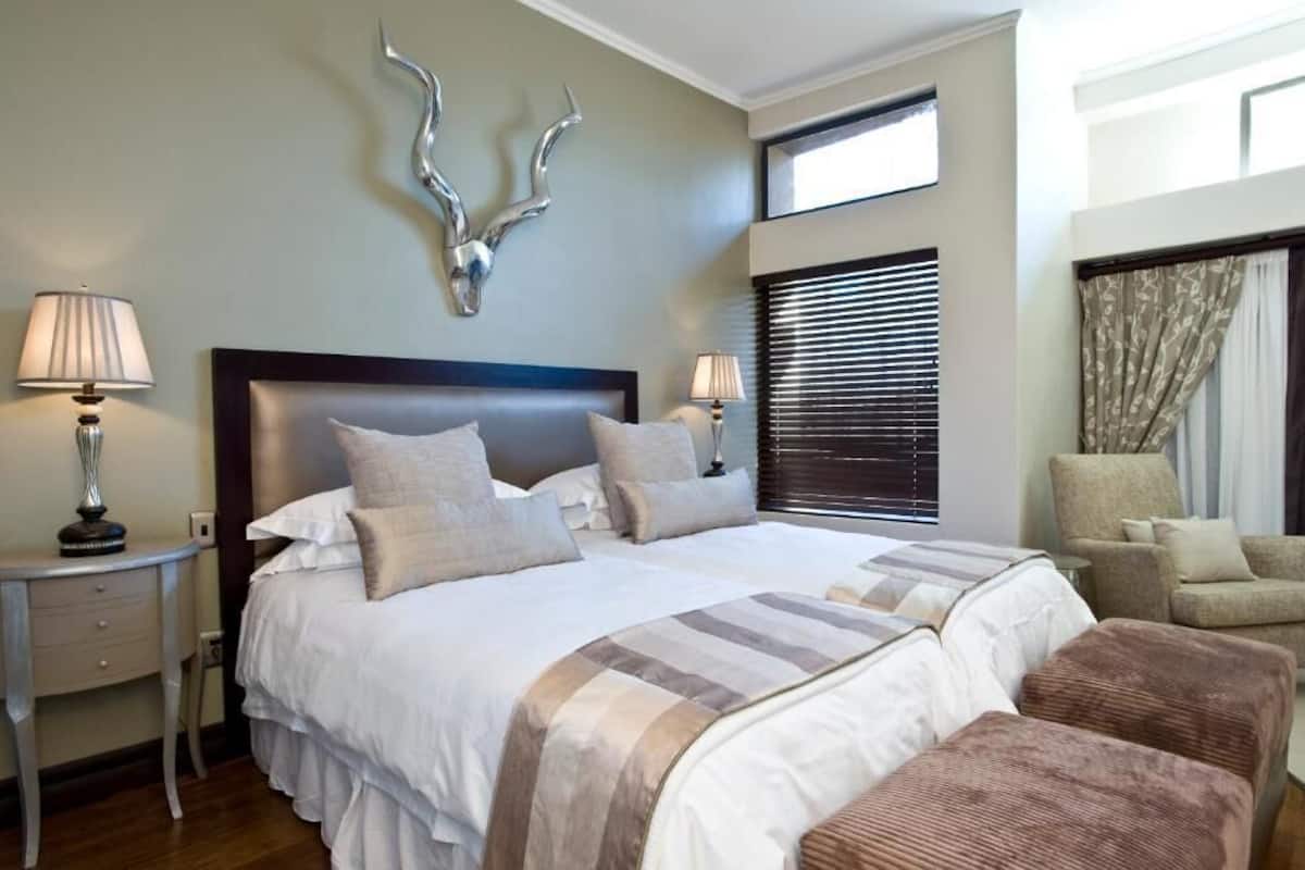A hotel room with a large bed, two bedside tables with lamps, a decorative wall piece, a window with blinds, and a chair.