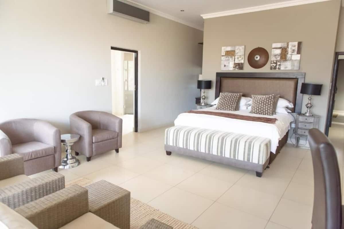 A spacious bedroom with a large bed, two armchairs, a small table, and a bedside table with lamps.