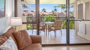 A balcony with wicker furniture, a glass door, and a view of palm trees and buildings. - Kamaole Sands 3-308, Premier Location, Renovated, Large Lanai, Wi-Fi, Split AC!  (Kihei)