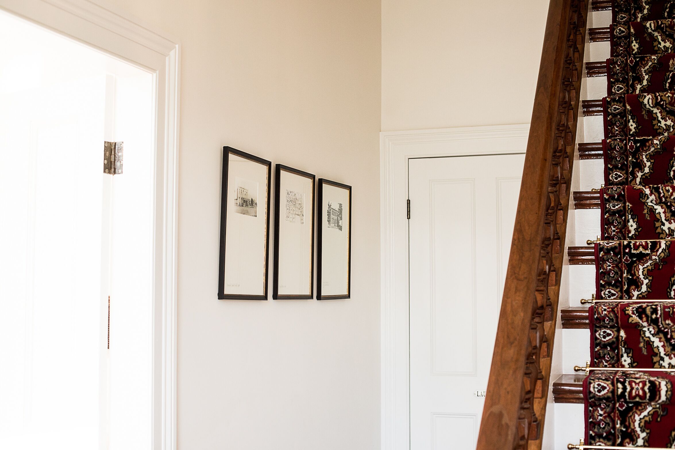 Un pasillo con una puerta blanca, tres cuadros enmarcados en la pared y una escalera de madera con tela estampada.