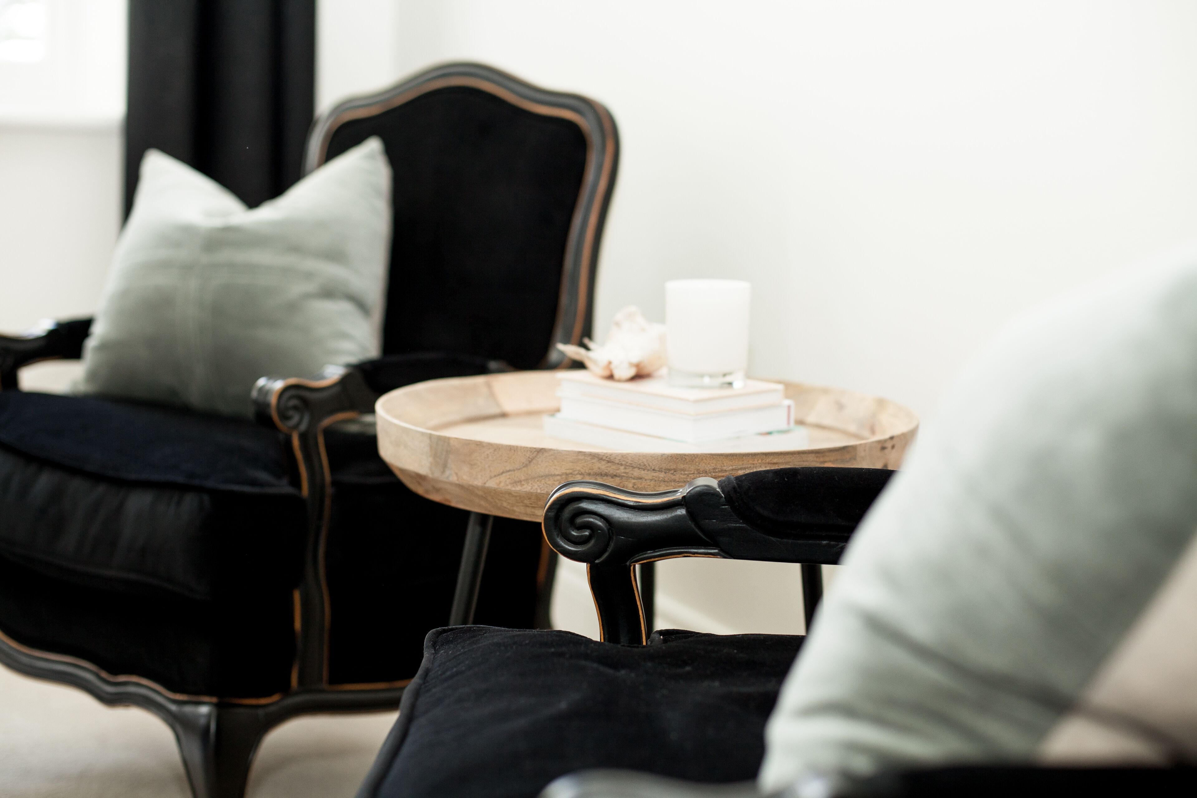A room with a black upholstered chair, a round wooden table with a white candle and books, and a light grey pillow.