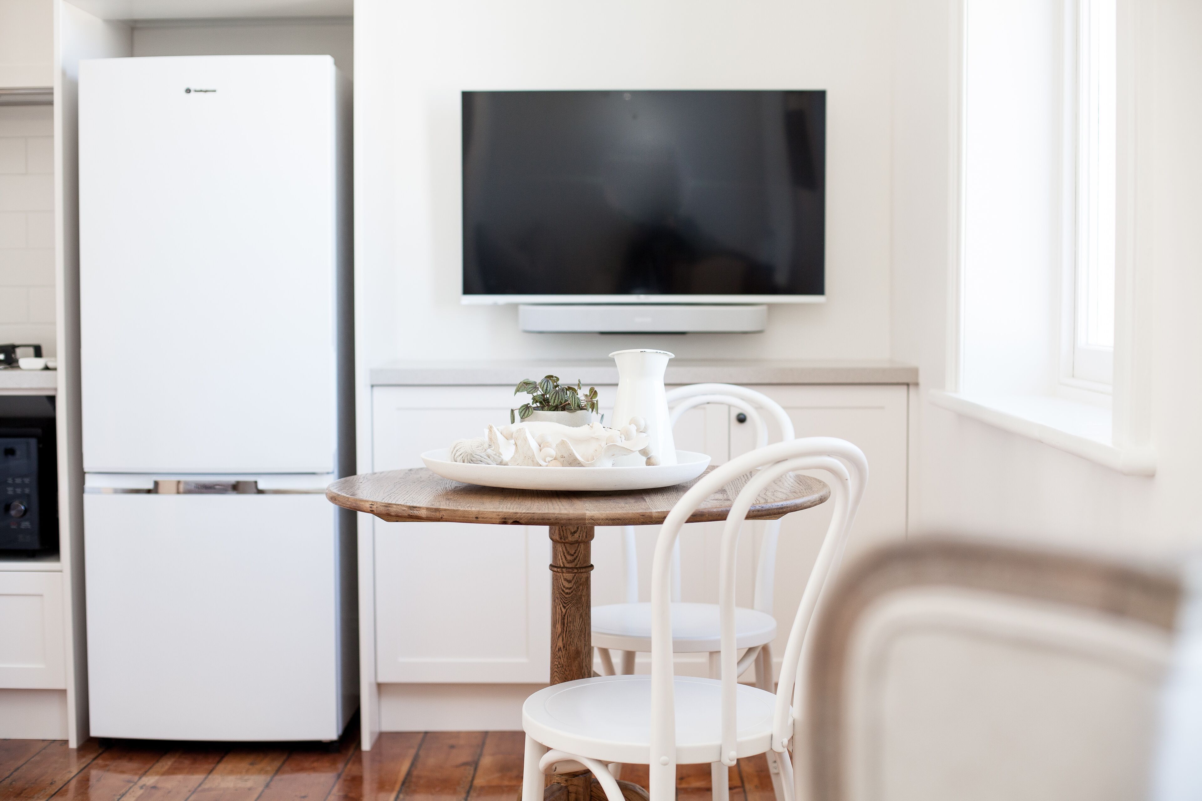 A modern kitchen with a white refrigerator, a flat-screen TV mounted above a wooden table, and white chairs.