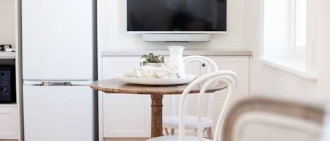 A modern kitchen with a white refrigerator, a flat-screen TV mounted above a wooden table, and white chairs.