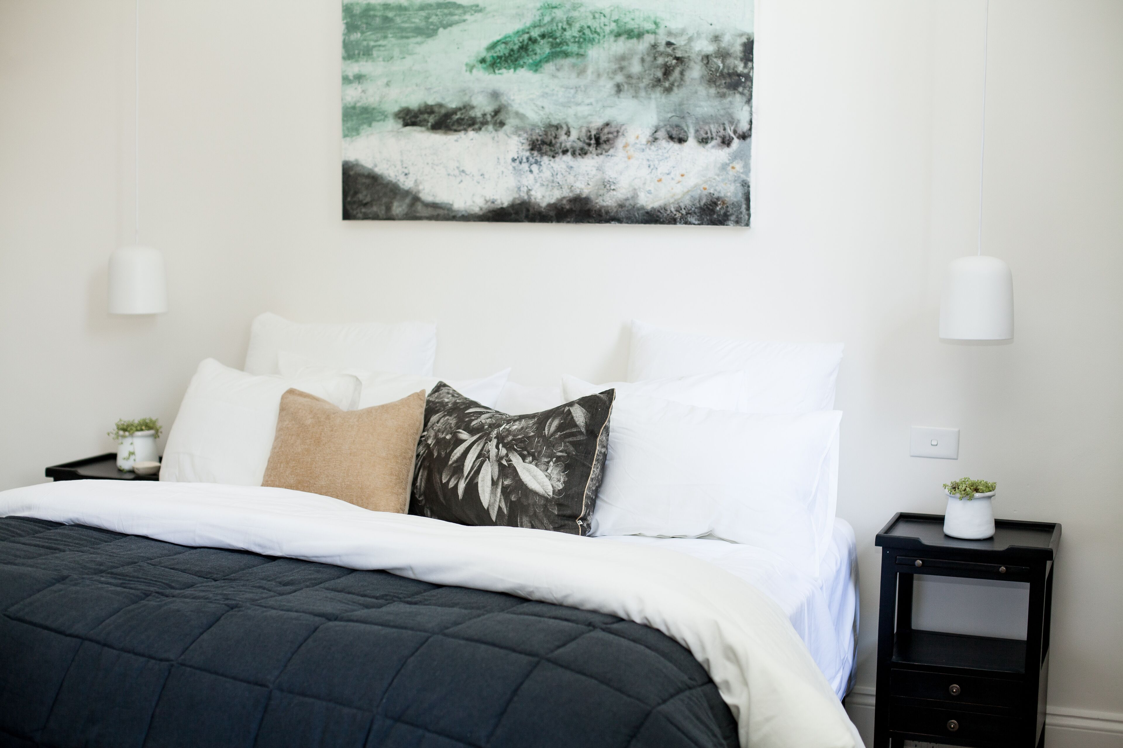 Uma cama bem arrumada com um edredom escuro, almofadas brancas e uma única almofada decorativa. Uma pequena planta está sobre uma mesa de cabeceira, e um quadro com uma paisagem natural está pendurado na parede.