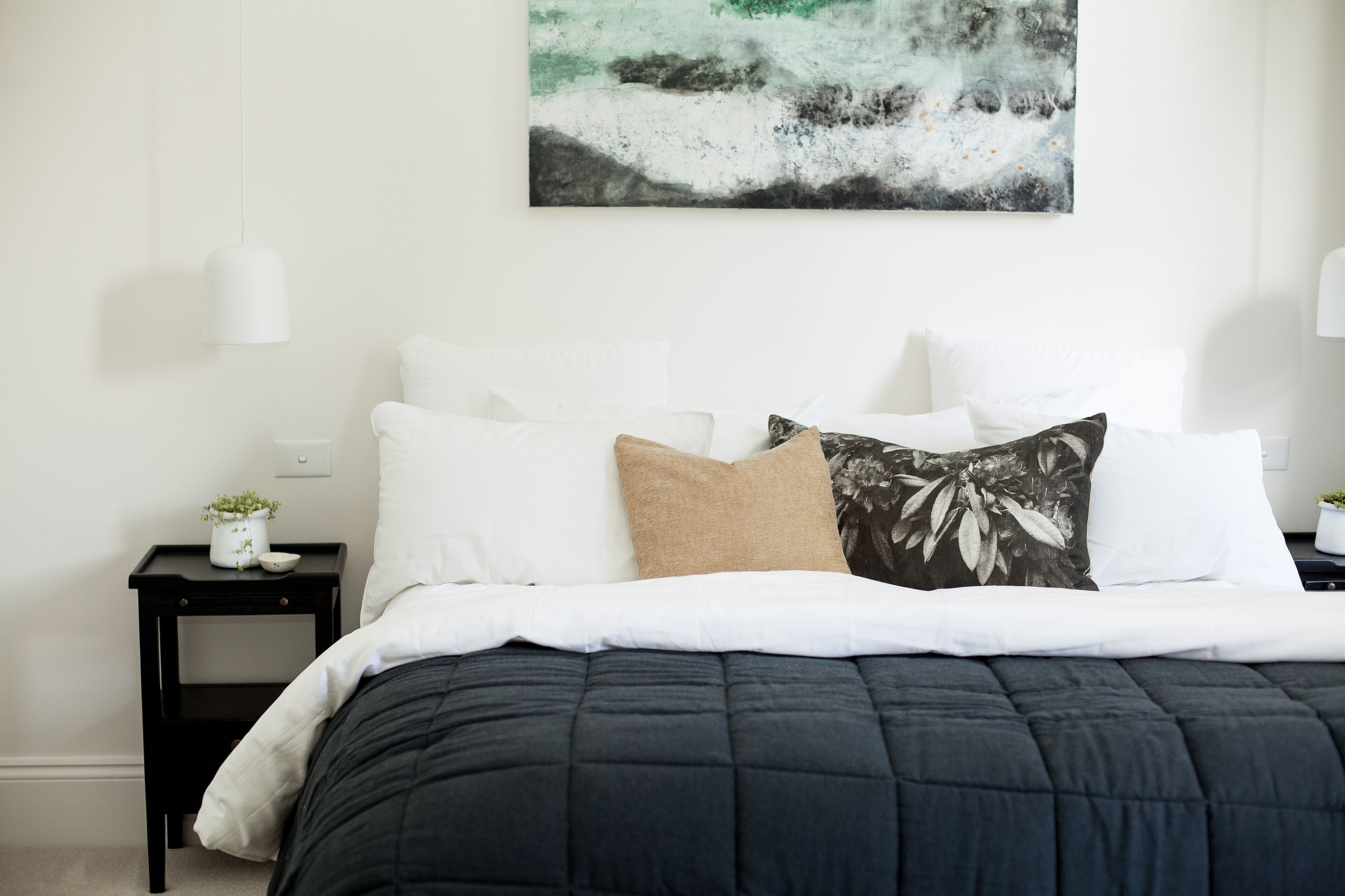 A neatly made bed with a dark quilt, white pillows, and a single decorative pillow. A small bedside table with a plant and a lamp is visible.