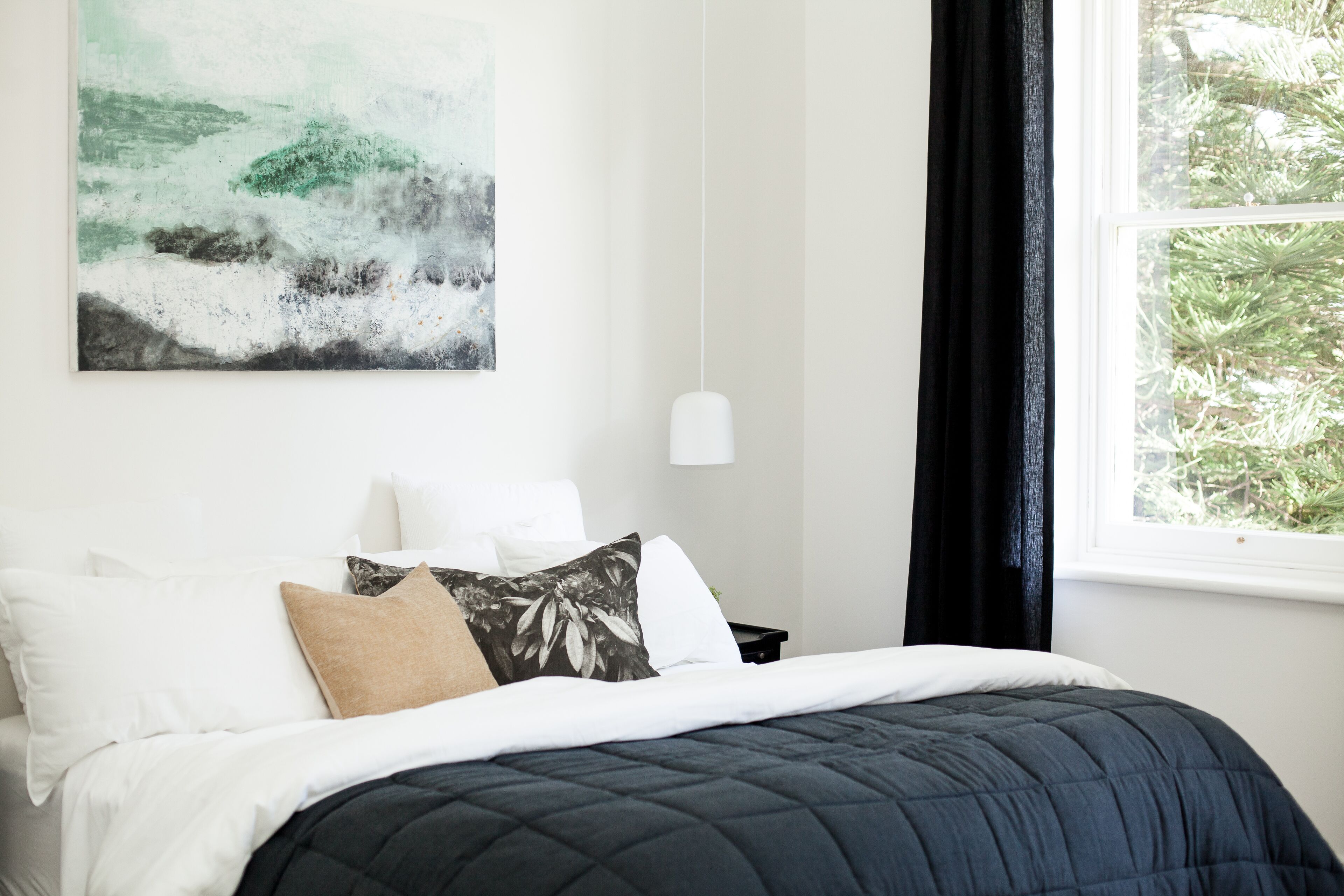 A bedroom with a bed, pillows, a painting, a nightstand, and a window with curtains.