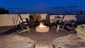 Una chimenea circular rodeada de asientos, ubicada en un patio empedrado con vista a un paisaje urbano al atardecer.