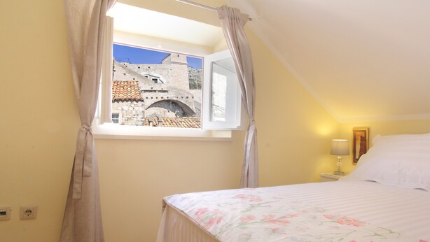 Un dormitorio con cama, ventana con cortinas, mesita de noche con lámpara y vista a un puente de piedra.