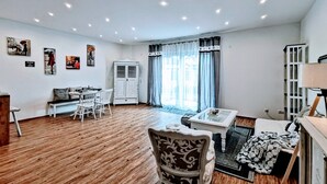 A spacious living room with wooden flooring, a dining area with white chairs, a white cabinet, and artwork on the walls.