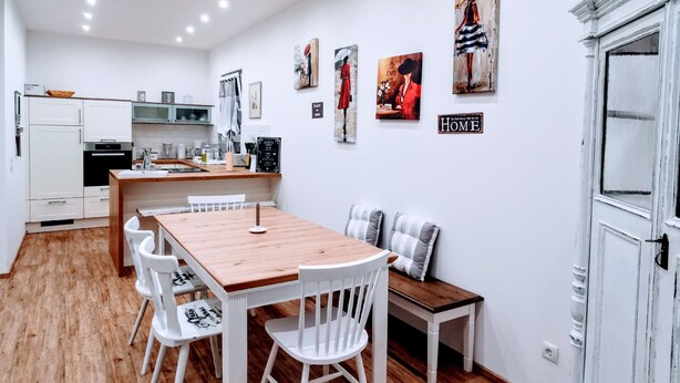 A modern kitchen with white cabinetry, a wooden dining table, and a bench. There are wall-mounted artworks and a sign that reads "HOME".