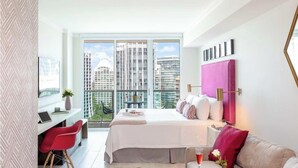 A modern bedroom with a large bed, a desk, and a view of tall buildings.