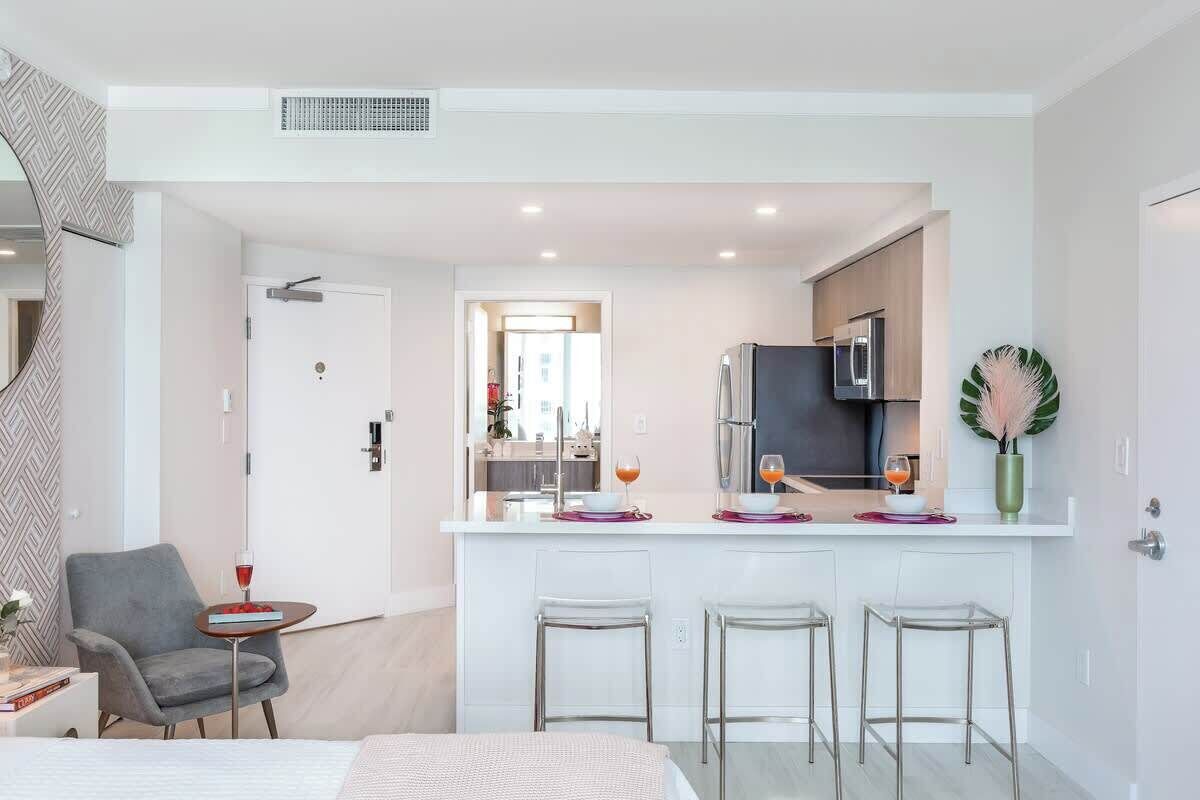 Una habitación de hotel moderna con kitchenette, una zona de comedor con taburetes de bar, un sillón gris y una mesita con una bebida.
