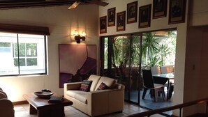 A living room with a sofa, a coffee table, and a dining area with chairs and a table. There are framed pictures on the wall and a ceiling fan.