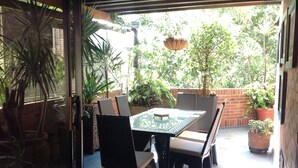An outdoor patio with a glass table and wicker chairs, surrounded by potted plants and a brick wall.