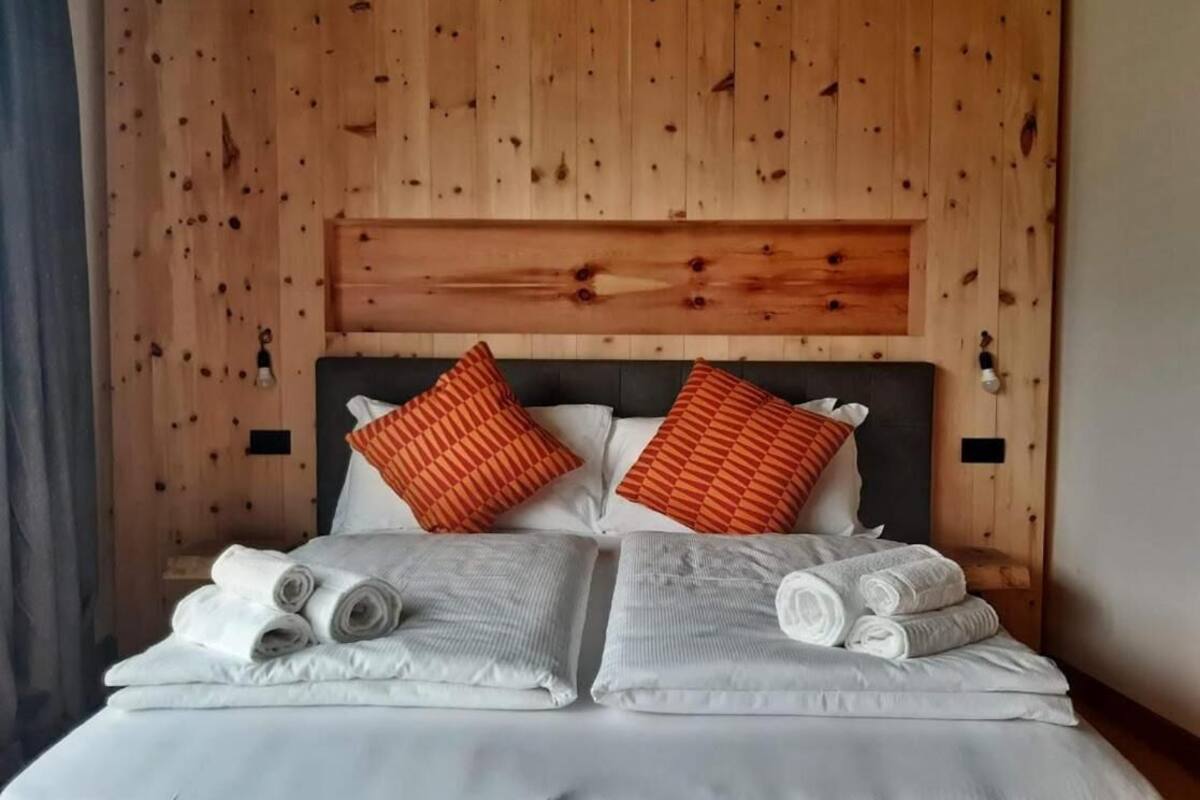 A neatly made bed with white linens and rolled towels, a wooden headboard, and two orange pillows.
