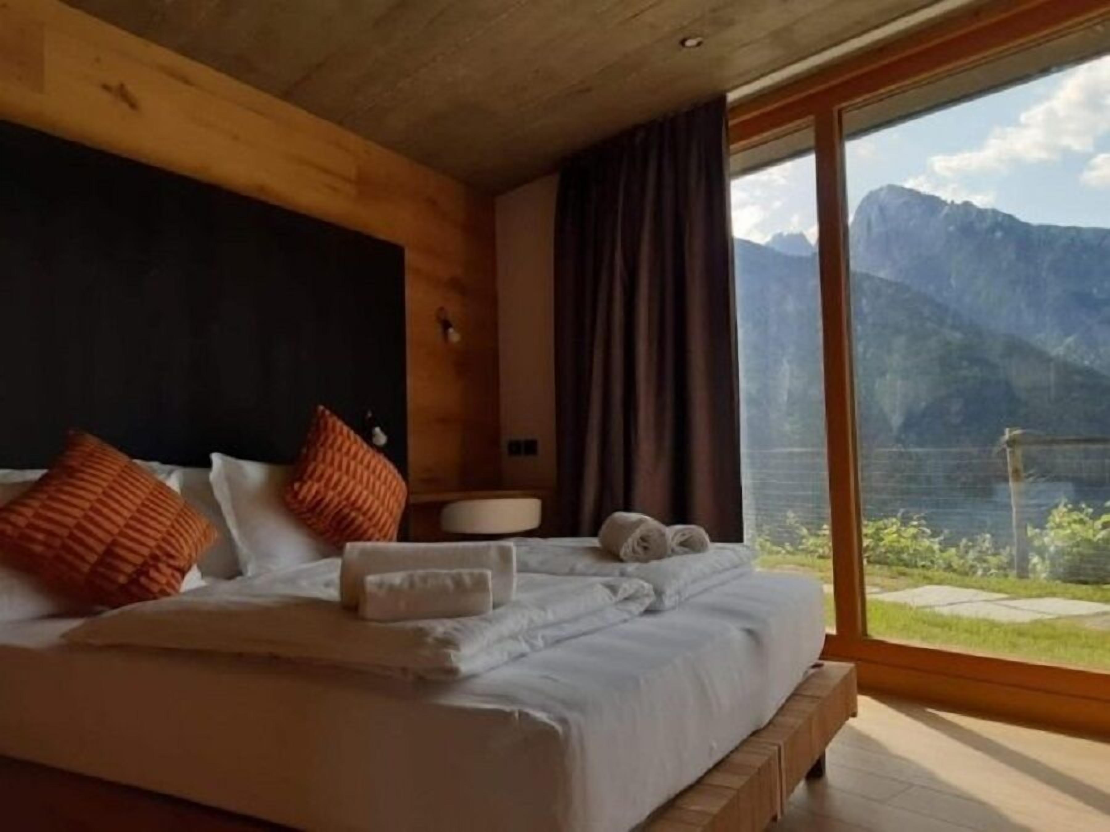Ein Schlafzimmer mit einem hölzernen Kopfende, einem großen Bett mit weißer Bettwäsche, zwei orangen Kissen, einem Nachttisch mit einer Lampe und Blick auf Berge und einen See durch ein großes Fenster.