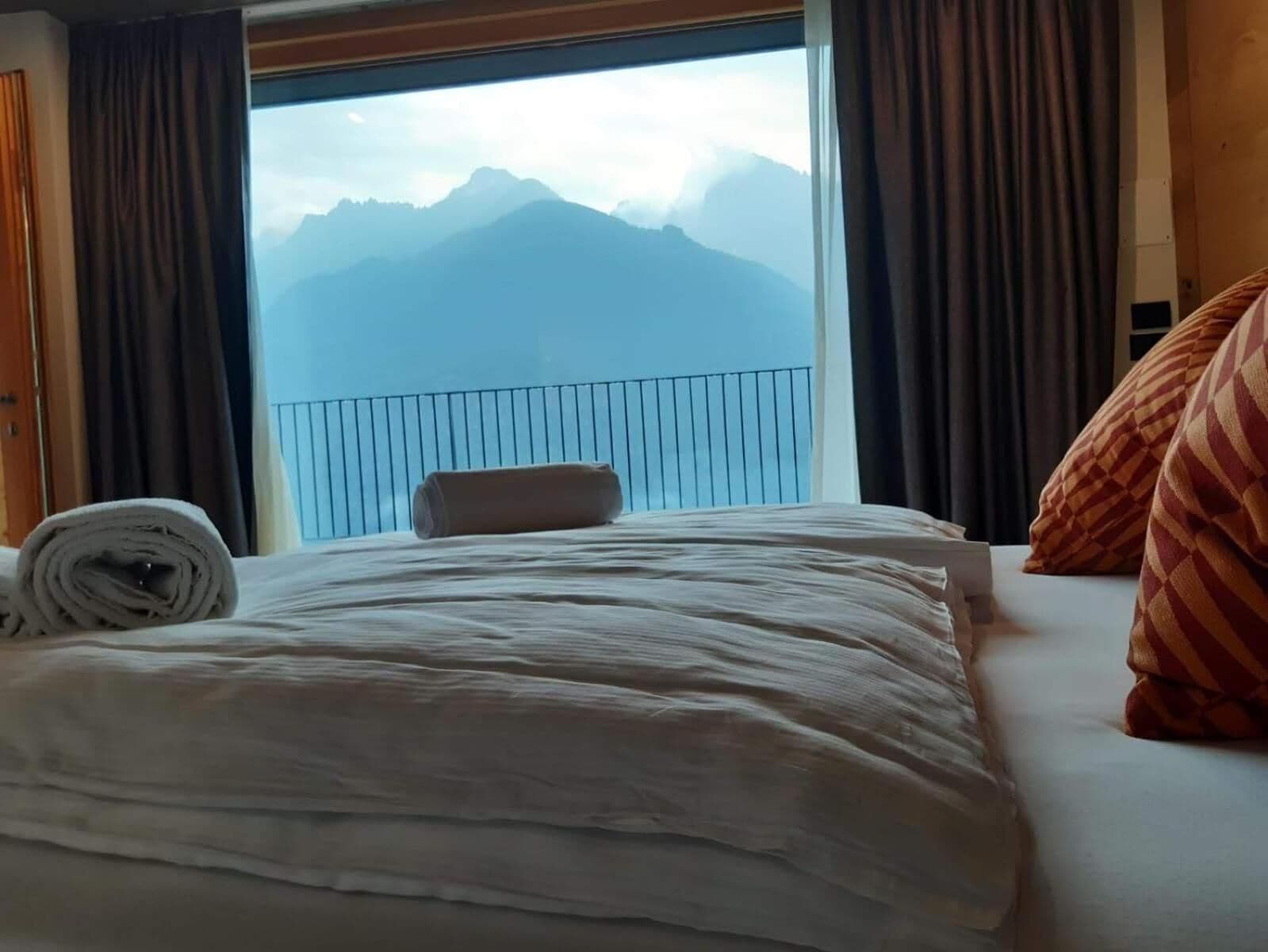 Une chambre d’hôtel avec un lit, une vue sur les montagnes par une grande fenêtre et une balustrade de balcon.