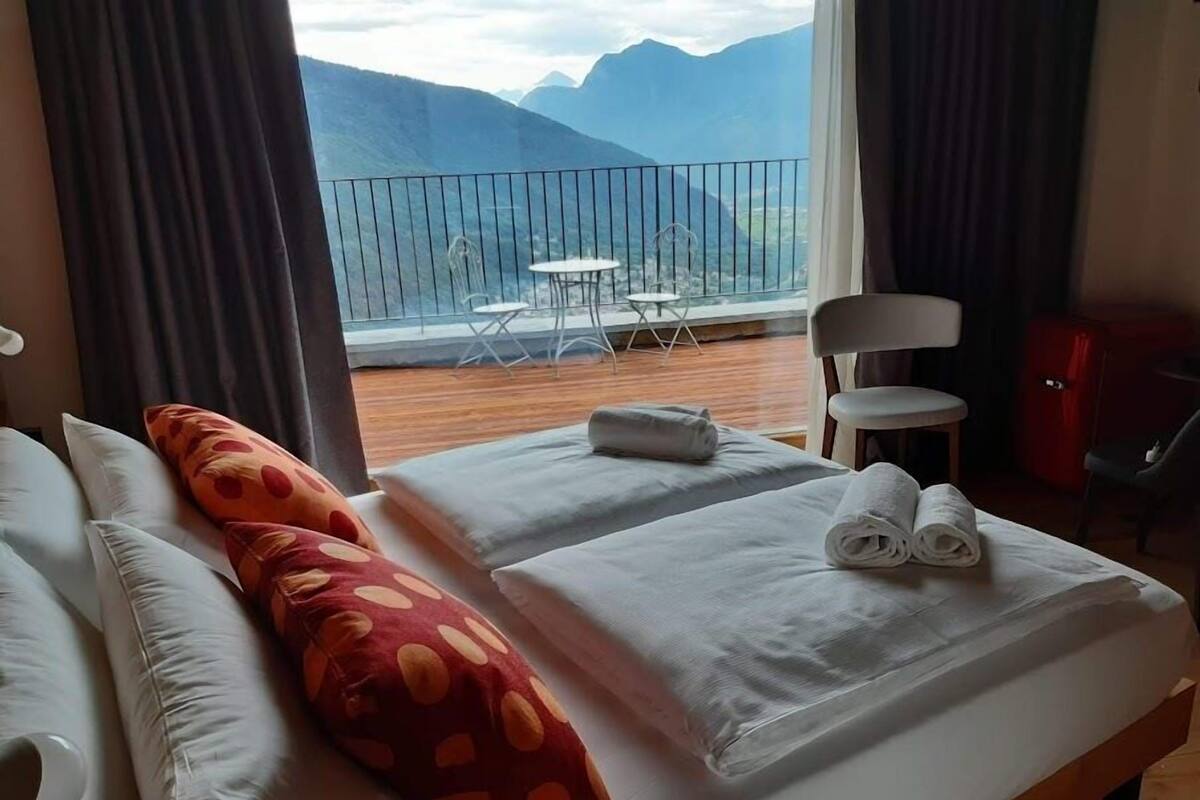 A hotel room with a bed, pillows, a small table, and a view of mountains through a balcony.