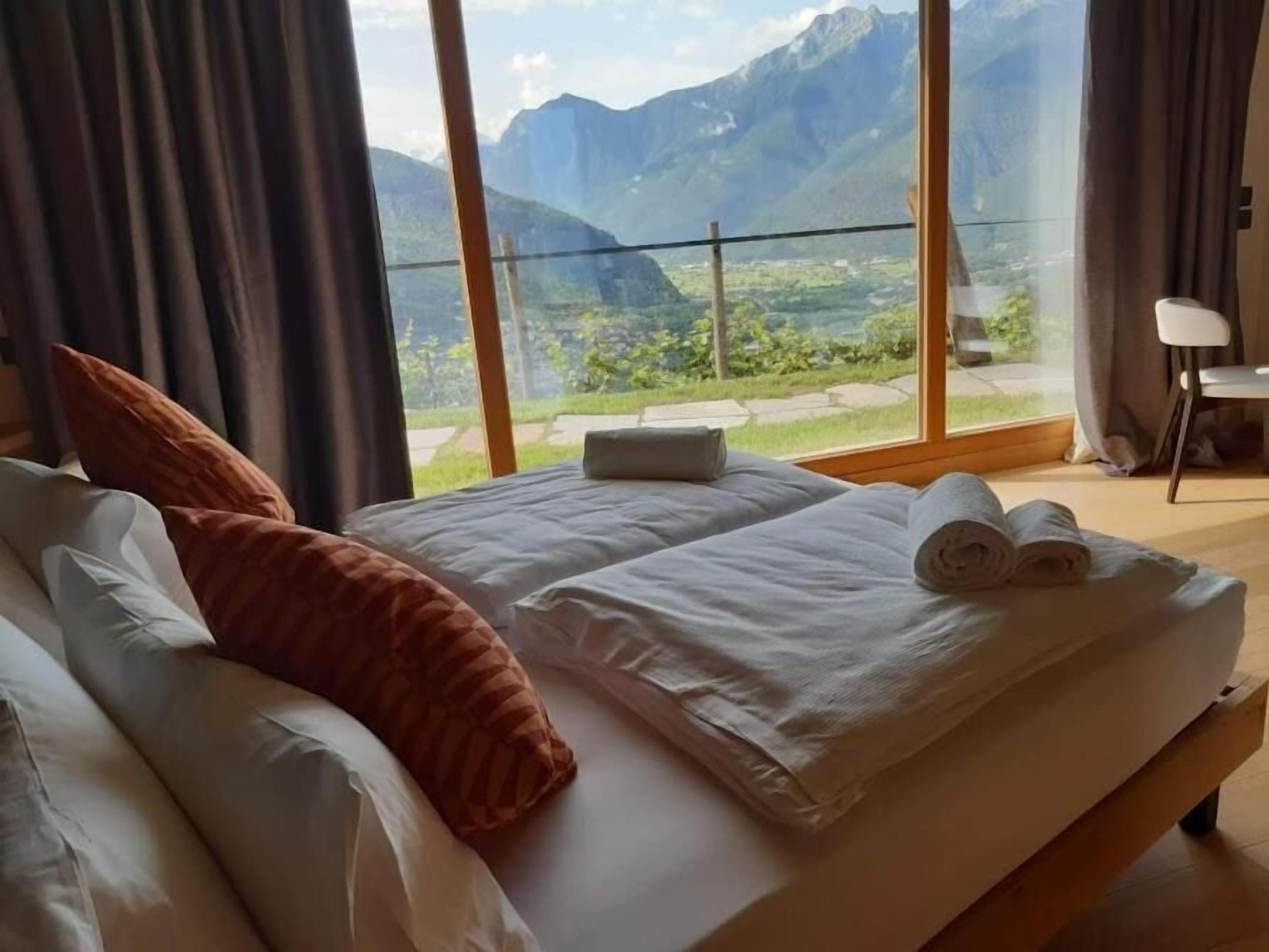 A hotel room with a bed, pillows, and a view of mountains through large windows.