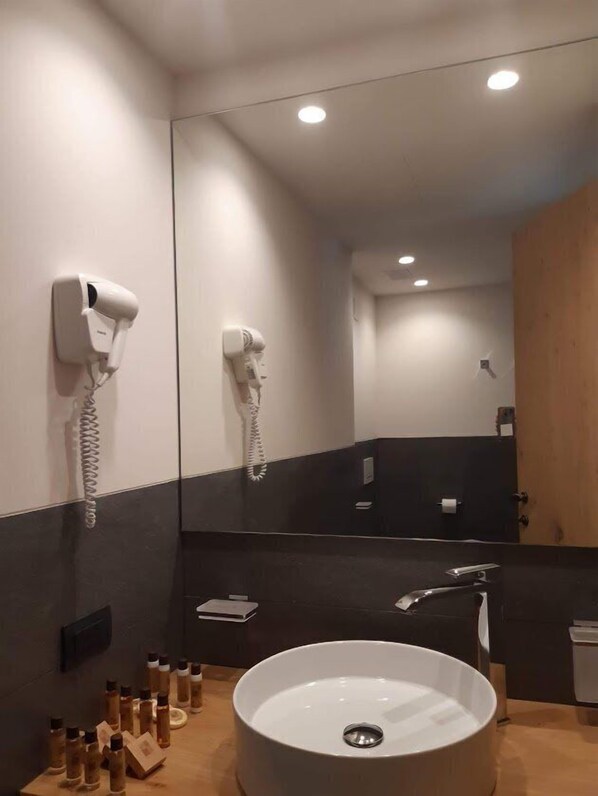 Ein modernes Badezimmer mit einem großen Spiegel, zwei an der Wand montierten Haartrocknern, einem runden Waschbecken und verschiedenen Pflegeprodukten auf der Ablagefläche.