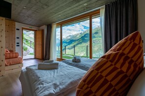 Ein Schlafzimmer mit großem Fenster und Blick auf die Berge, einem Bett mit weißer Bettwäsche und einer hölzernen Bank mit Kissen.