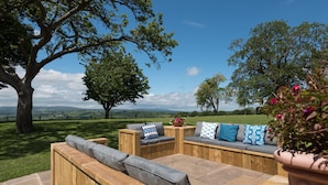 Une terrasse en bois avec des bancs intégrés, un grand arbre et une vue sur la campagne.