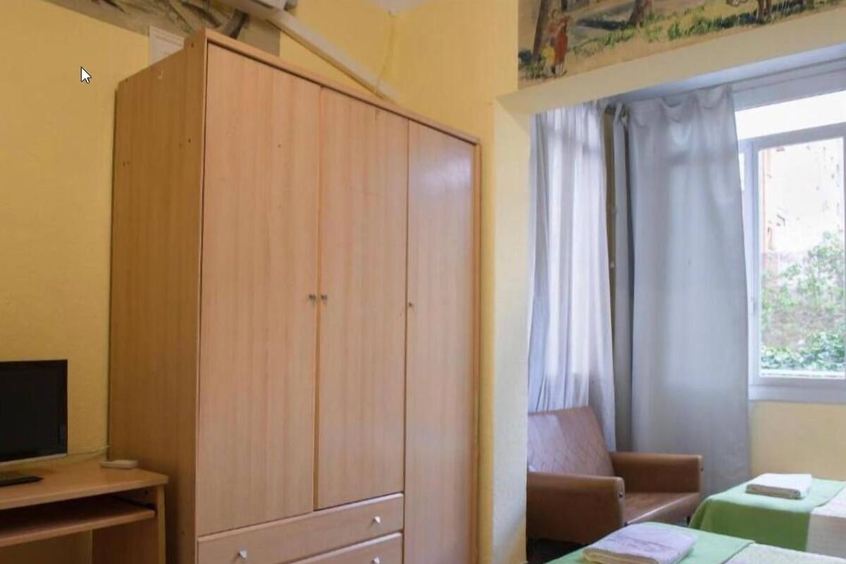 Una habitación con un armario de madera, un escritorio con computadora, dos camas verdes, una ventana con cortinas y un cuadro en la pared.