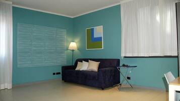 A room with teal walls, a dark blue sofa with two pillows, a lamp, a painting, and a window with white curtains.
