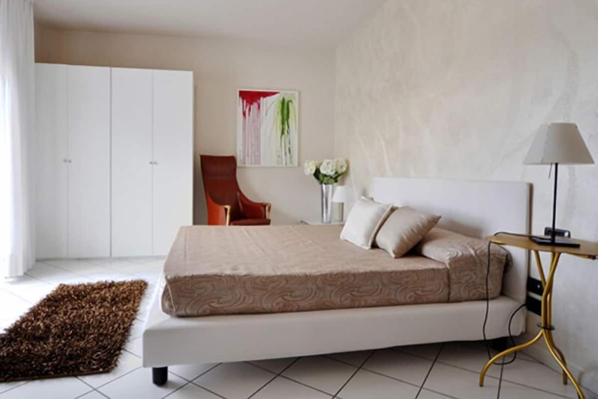 A bedroom with a bed, a chair, a bedside table with a lamp, a painting on the wall, and a rug on the floor.