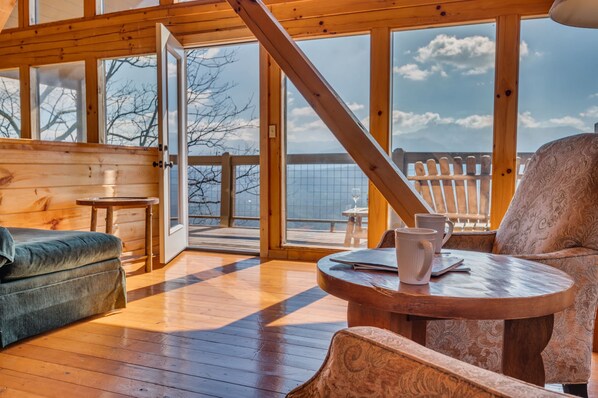 A wooden cabin interior with a sofa, a table with a mug, and a view of the outside.