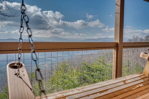 Une terrasse en bois avec une chaise suspendue, une rambarde en métal et une vue sur un paysage montagneux.